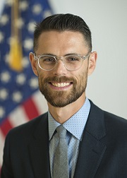 Dr. Ostrovsky wearing a blue collared shirt, grey tie and dark jacket in front of an American flag