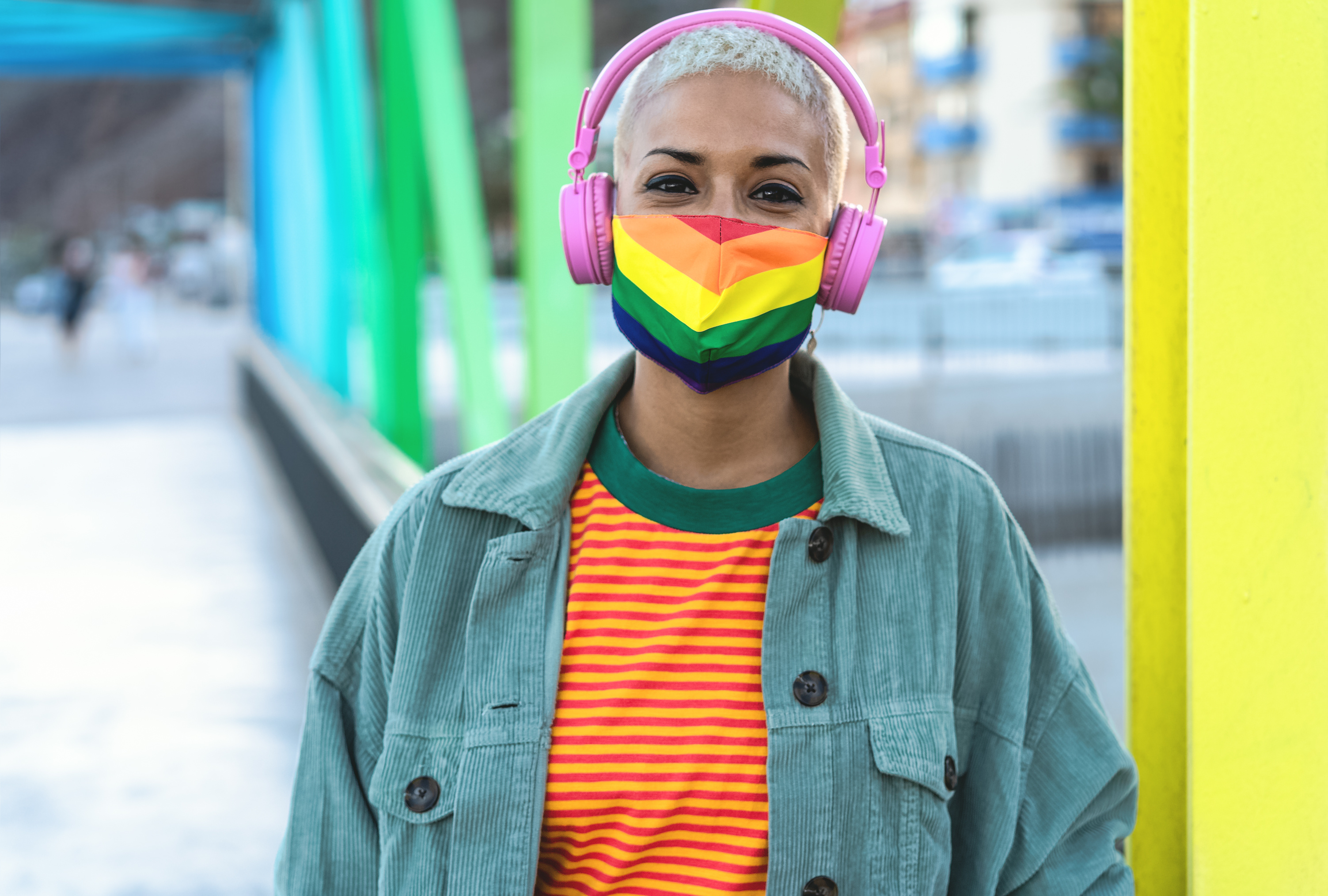 A black individual in a teal corduroy jacket, orange and yellow striped shirt with a green collar, with a rainbow face mask and pink headphones