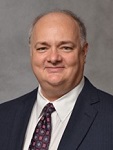 This is an image of Dr. Paul Orchard. He is wearing a dark navy-blue suit and coordinating patterned tie. He has a slight smile.