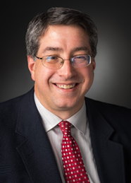 This is a photo of Dr. Daniel DeAngelo. He is facing the camera and smiling and wearing glasses, a dark suite and red patterned tie. 