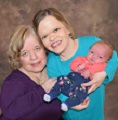 Grandma Deb is on the left handside wearing a purple shirt, Daugther Maria who is holding Baby Rolland is wearing a teal shirt. All members of the Hecht family are facing the camera smiling. Loving potrait of a little family.