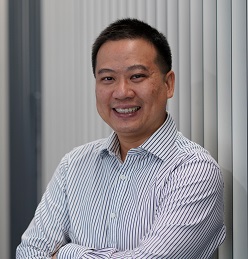 Dr. Jeremy Lim is standing in front of vertical white blinds, facing the camera smiling and wearing a white button down shirt with small blue stripes.