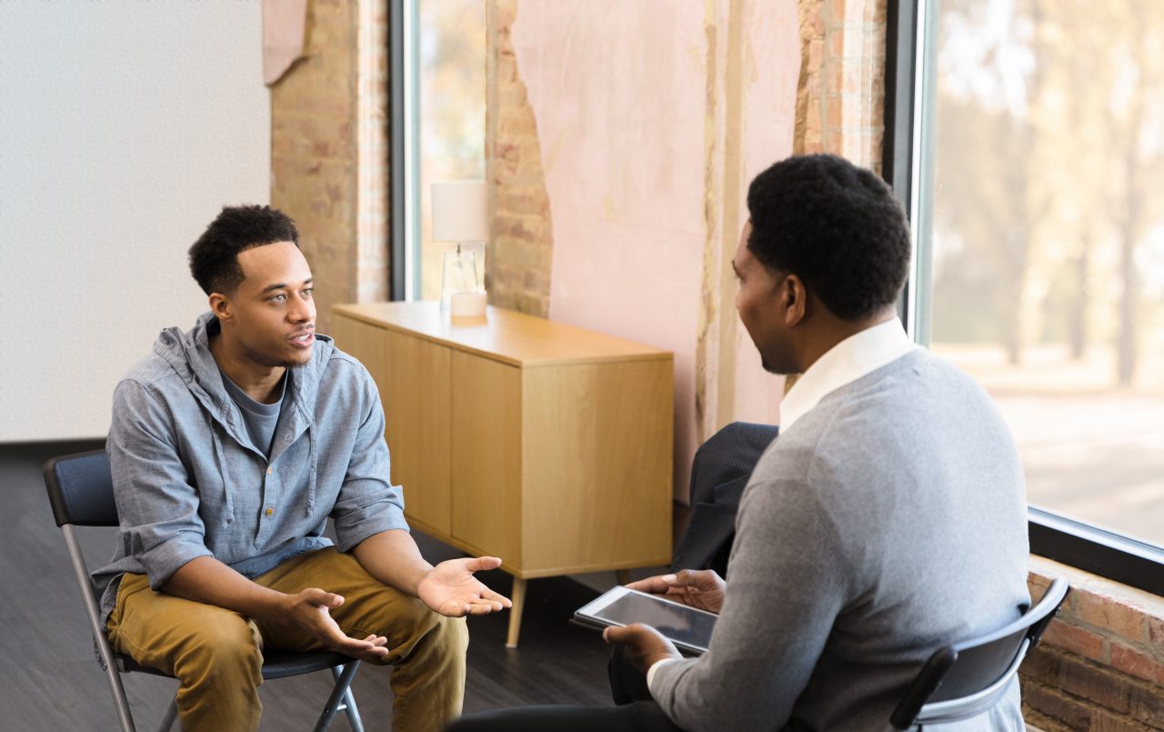 A Black man with tan pants and a grey shirt sits opposite a Black man in black pants, a white collared shirt and a grey sweater holding an ipad