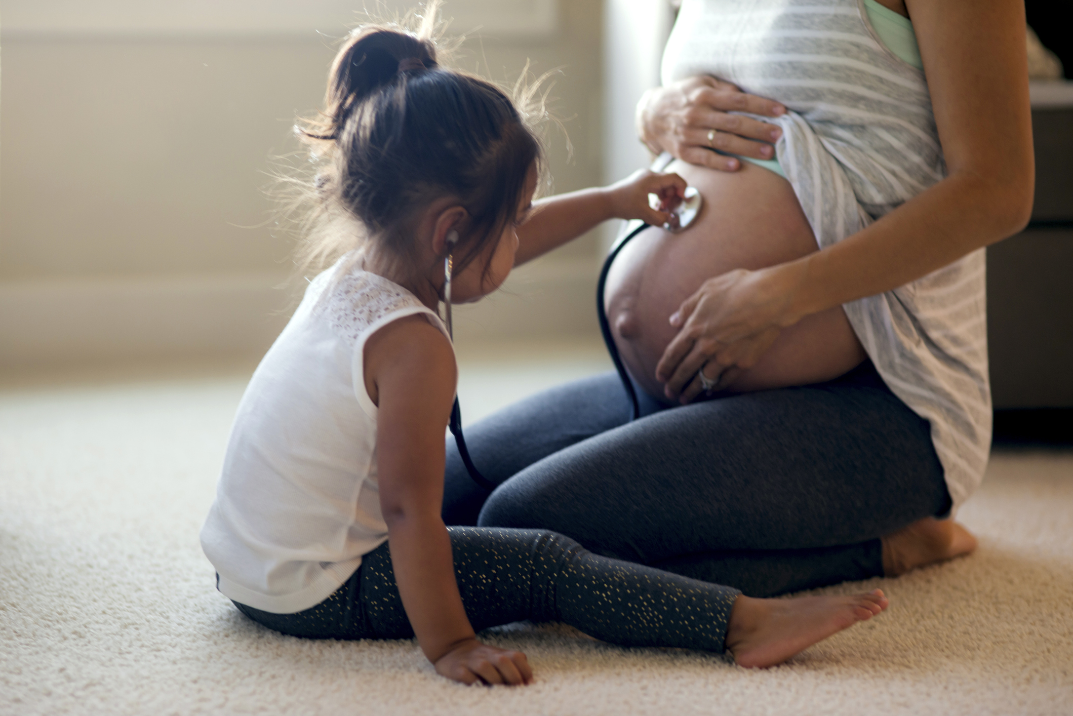 A young girl holds a stethoscope to a pregnancy woman's belly