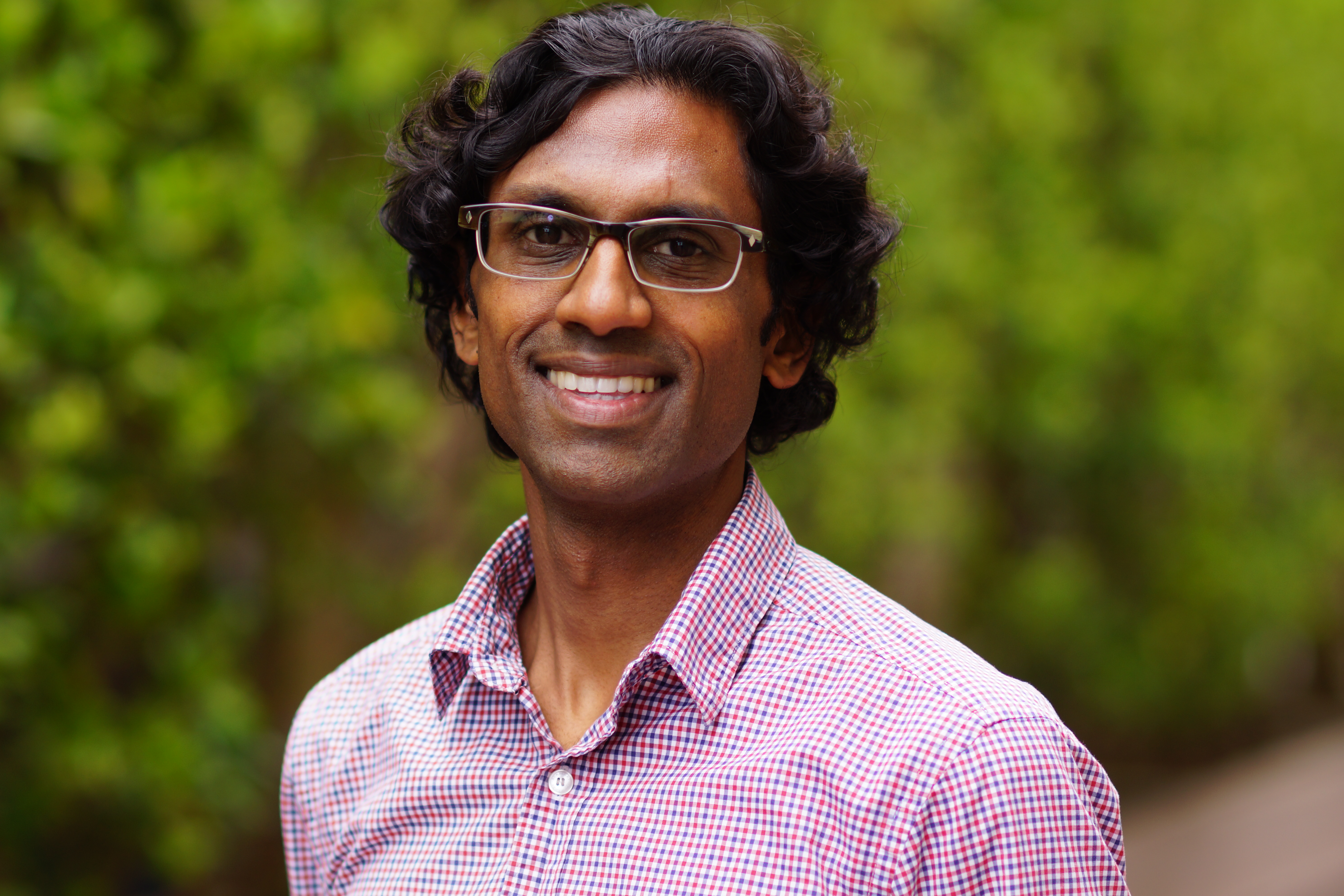 Dr. Nirav Bhakta wearing a pink shirt and glasses standing in front of foliage