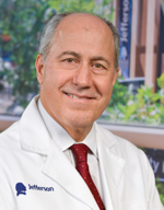 Dr. Karalis is an expert in the field of cardiology and lipidology. He is facing the camera and slightly smiling. He is balding and has grey hair. He is wearing a white lab coat, white dress shirt and red tie. 