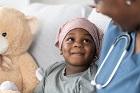 young African American girl in a bed with her teddy bear next to her, a doctor or nurse in blue scrubs with a stethoscope is next her; they are both smiling 