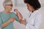 Woman in green top with glasses and grey hair showing her irration/rash on her arm to her health care professional. Health care professional is woman wearing a white lab coat.