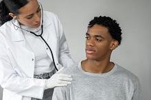 A black woman in a white coat and white gloves listens to a stethoscope which is placed on the back of a black man with short hair wearing a grey sweatshirt