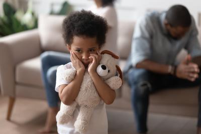 A young black child holding a white stuffed animal holds their hands to their face in distress while a couple sits behind them